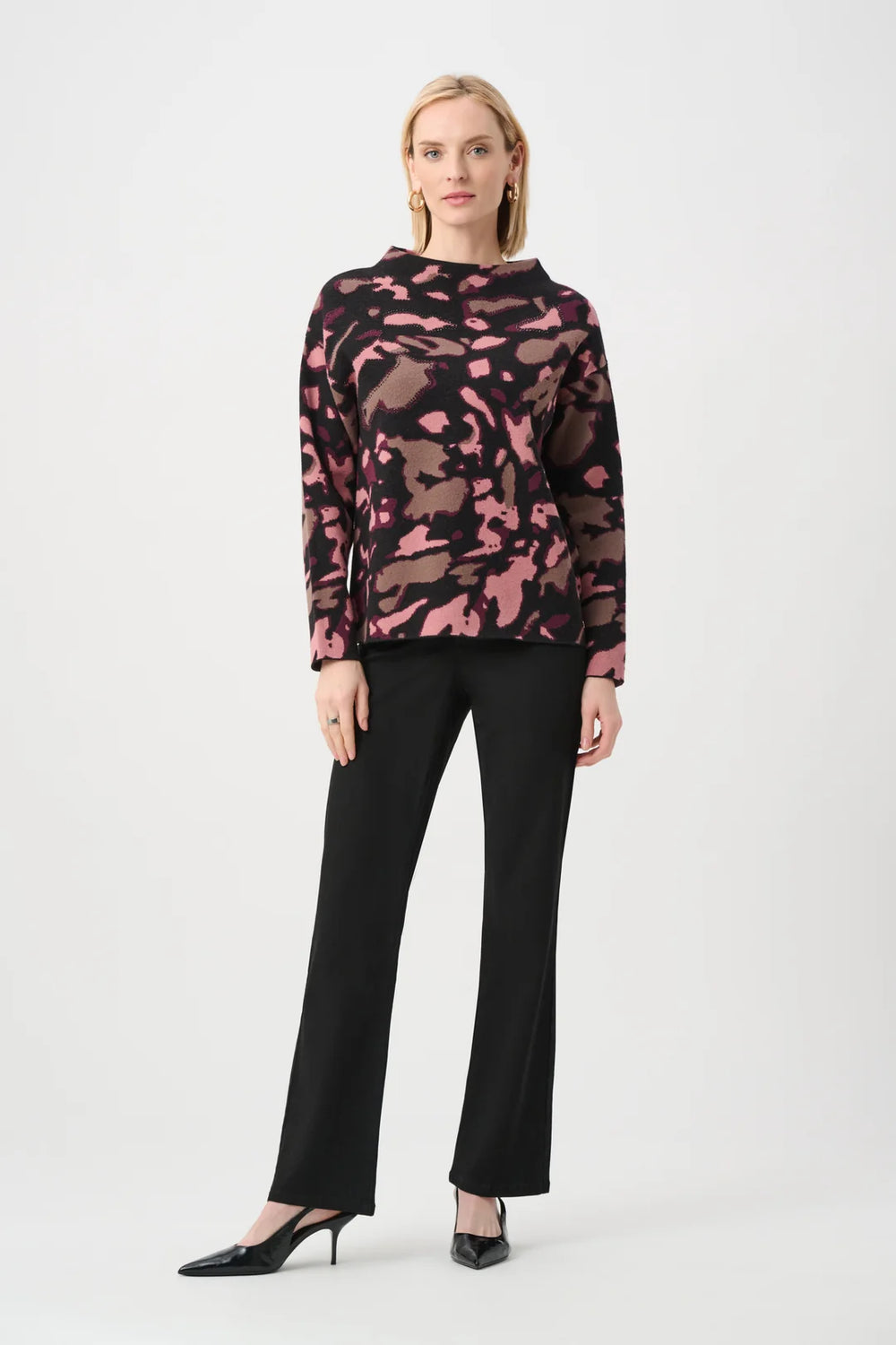 A woman modeling Joseph Ribkoff Abstract Print Sweater Style 263930, a long-sleeve mock-neck top with a pink and black abstract camouflage pattern, paired with black trousers and black pointed-toe heels, against a light gray studio background.