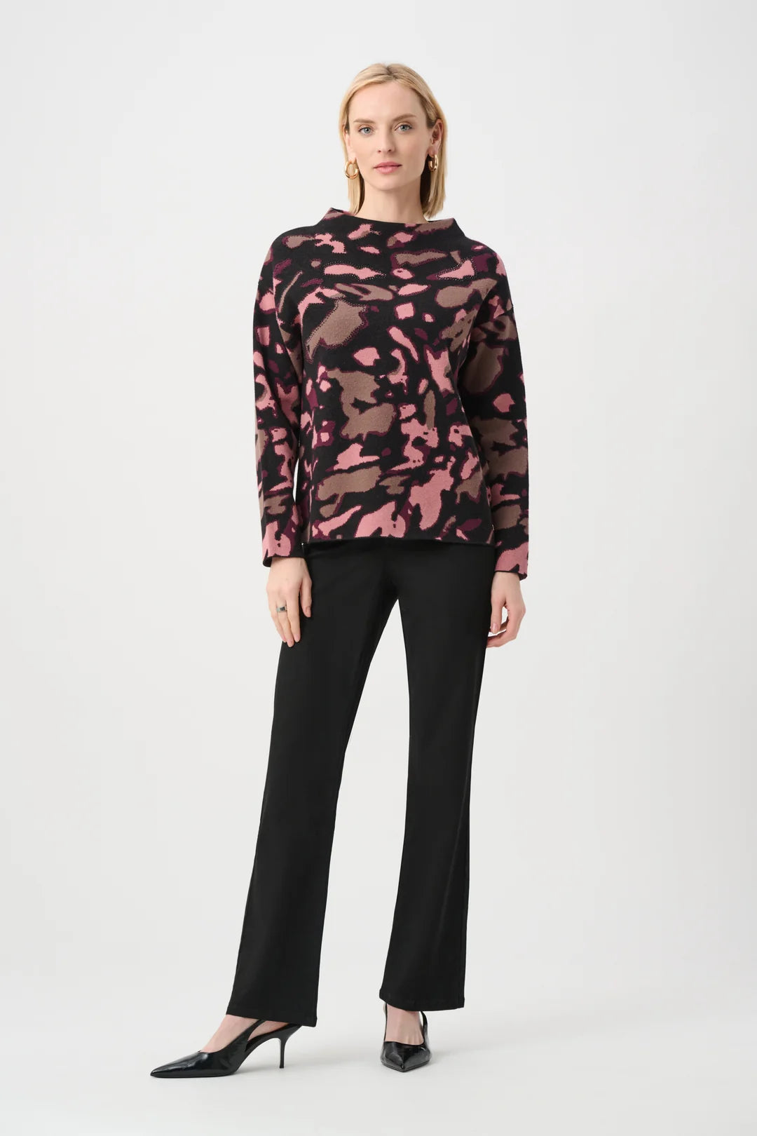 A woman modeling Joseph Ribkoff Abstract Print Sweater Style 263930, a long-sleeve mock-neck top with a pink and black abstract camouflage pattern, paired with black trousers and black pointed-toe heels, against a light gray studio background.
