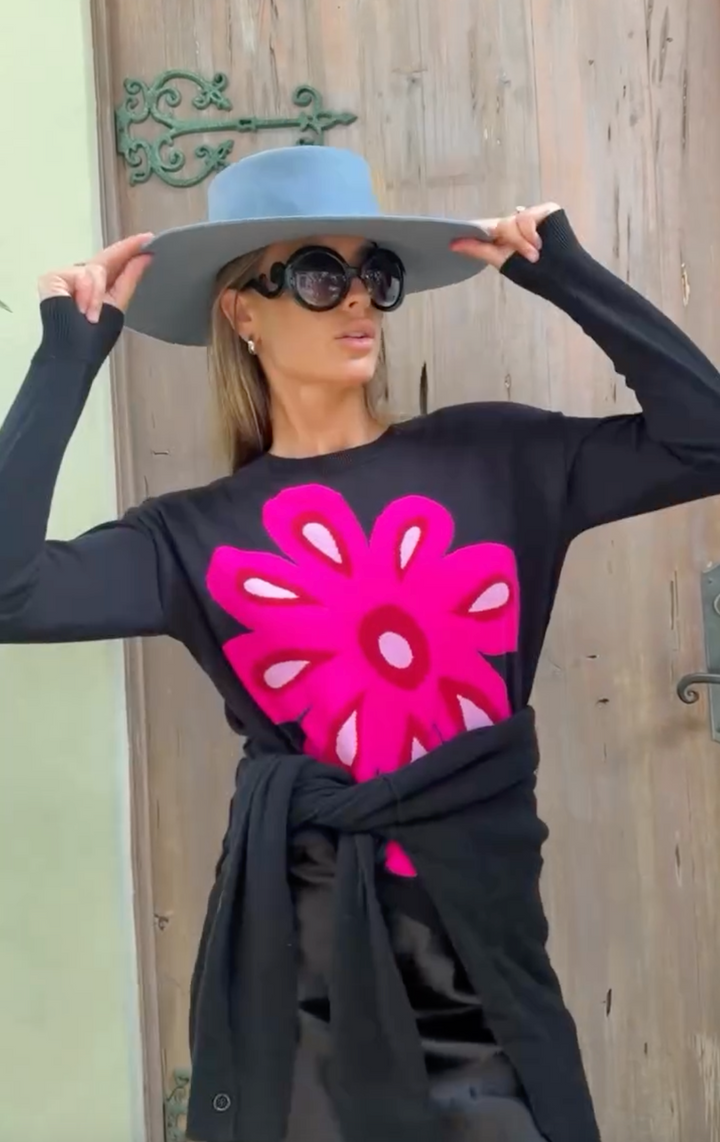 A Women in sunglasses wearing a black Lisa Todd Flower Power Cotton Sweater, striking a pose with her hands on her light blue bolero hat in front of a rustic wood door.