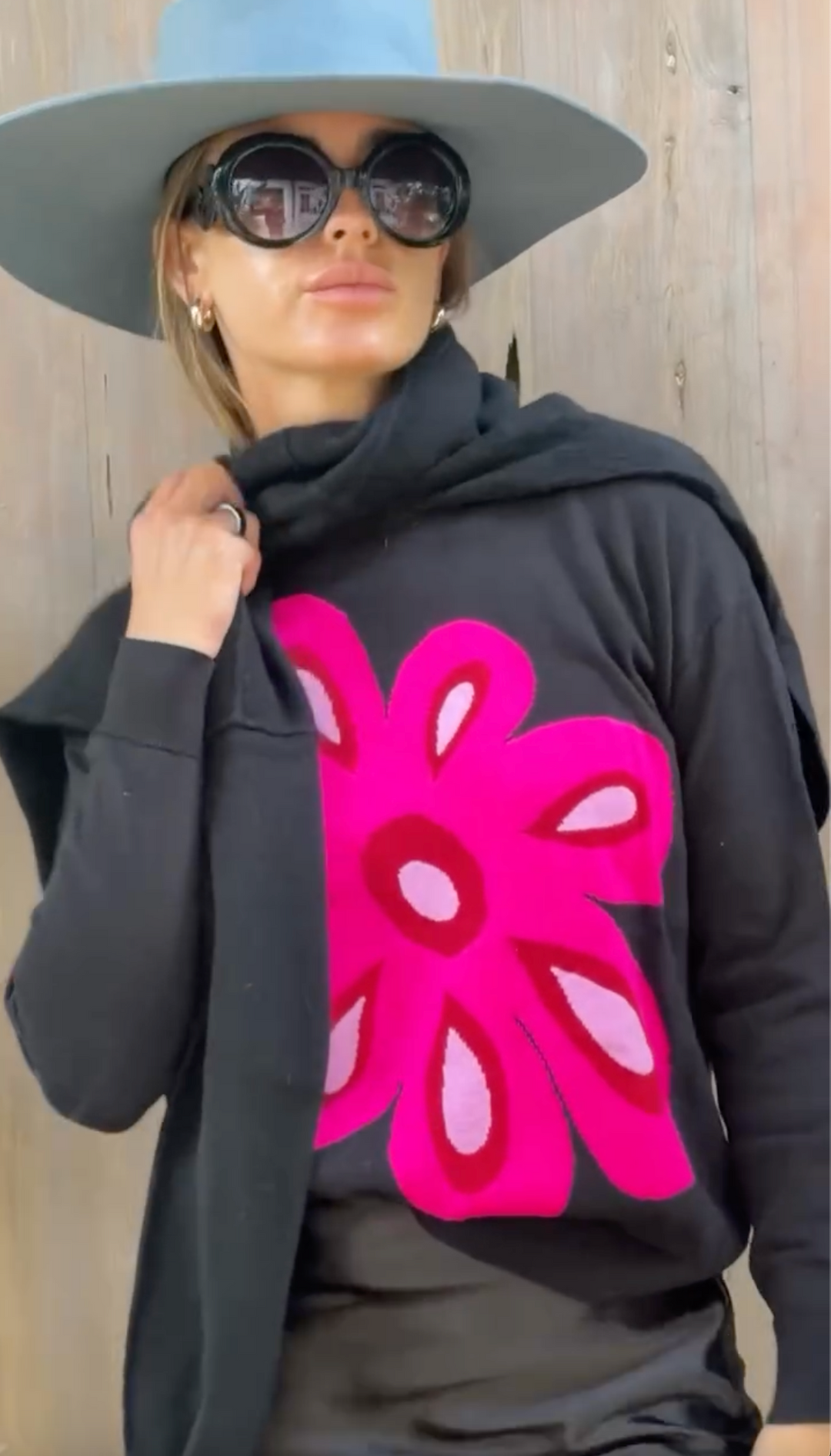 A close-up of a woman wearing a black a large bright pink Lisa Todd Flower Power Sweater styled with a wide-brimmed light blue bolero hat and dark sunglasses, standing against a light wood plank wall.