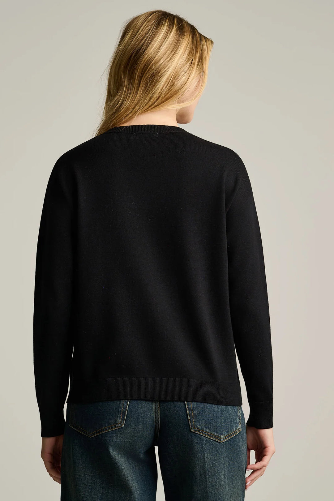 A woman viewed from the back wearing a black long-sleeve knit Lisa Todd Flower Power Sweater and blue jeans against a plain light gray studio background.
