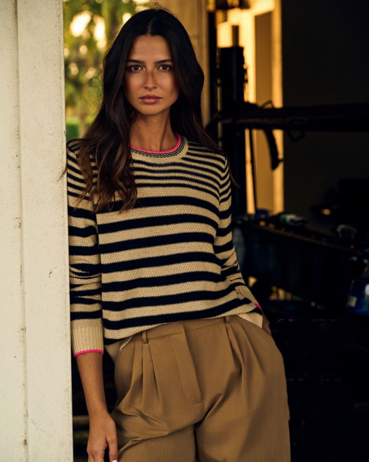 A woman wearing a Lisa Todd Stripe Idol Sweater with pink trim and tan trousers, leaning against a door frame in a dimly lit room.